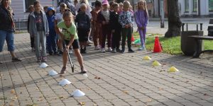 Bērni stāv divās kolonās, divi bērni priekšā vada bumbu gar konusiem ar florbola nūju.