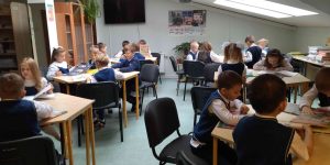 Bērni bibliotēkā sēž pie galdiem un skatās grāmatas