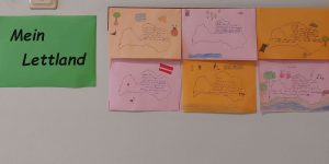 Plakāts ar tekstu "Mein Lettland" un blakus viņam bērnu veidotie tekstiņi vācu valodā kur aprastīta Latvija