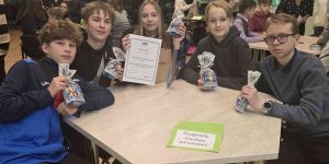 Bērni pie galda sēžot, rāda savus diplomu un konfektes.