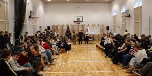 Bērni sēz kolonās sporta zāles malās un skatās priekšnesumus