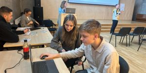 Skolēni strādā pie datora zālē kāmēr fonā vadītājs runā