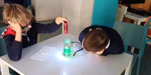 Bērns spīdina lāzeri glāzē ar ūdieni kāmēr otrs bērns novēro glāzi