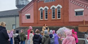 Bērni stāv priekšā sarkanai ķieģeļu ēkai un klausās gidu